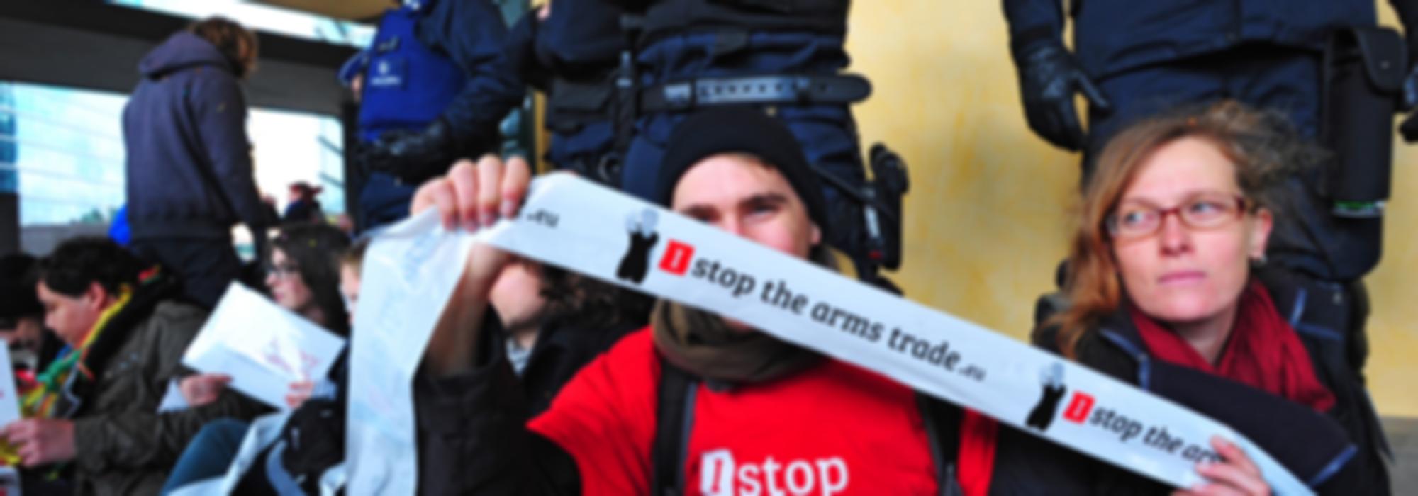 actiefoto met een aantal activisten aan een gebouw waarvoor de politie staat. Een activist houdt een lint met I Stop The Arms Trade vast. 