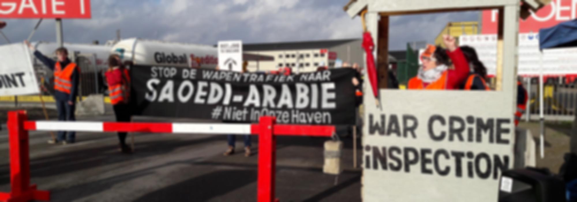 protestfoto bij de haven van Antwerpen (katoennatie) met grote  zwarte banner waarop witte letters staan die zeggen: "Stop de wapentrafiek naar Saoedi-Arabië" naast de banner staat een douanehuisje met slagboom, waarop staat "War Crime inspection" in het huisje staat een vrouw met een opgeheven vuist