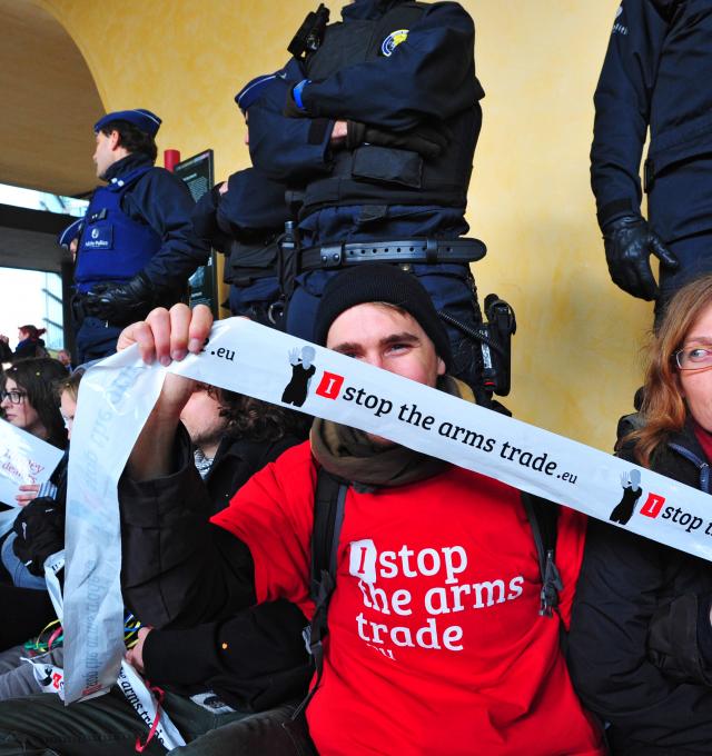 actiefoto met een aantal activisten aan een gebouw waarvoor de politie staat. Een activist houdt een lint met I Stop The Arms Trade vast. 