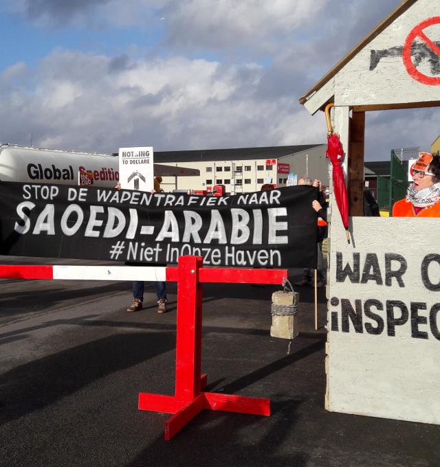 protestfoto bij de haven van Antwerpen (katoennatie) met grote  zwarte banner waarop witte letters staan die zeggen: "Stop de wapentrafiek naar Saoedi-Arabië" naast de banner staat een douanehuisje met slagboom, waarop staat "War Crime inspection" in het huisje staat een vrouw met een opgeheven vuist